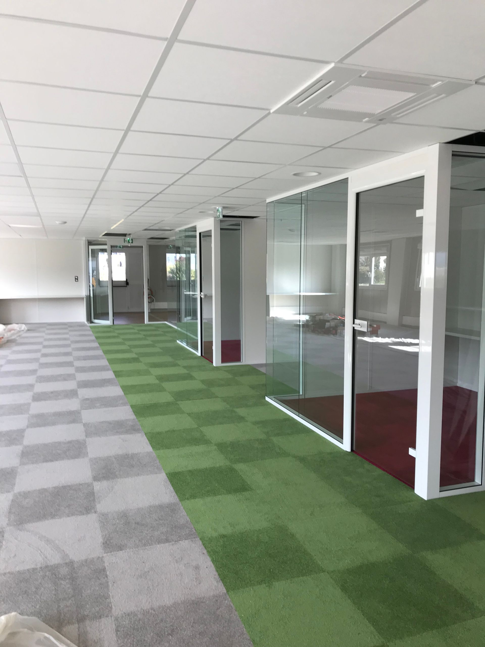 Bureau vide avec moquette à carreaux verts et gris, bureaux aux parois de verre et plafonds et murs blancs.