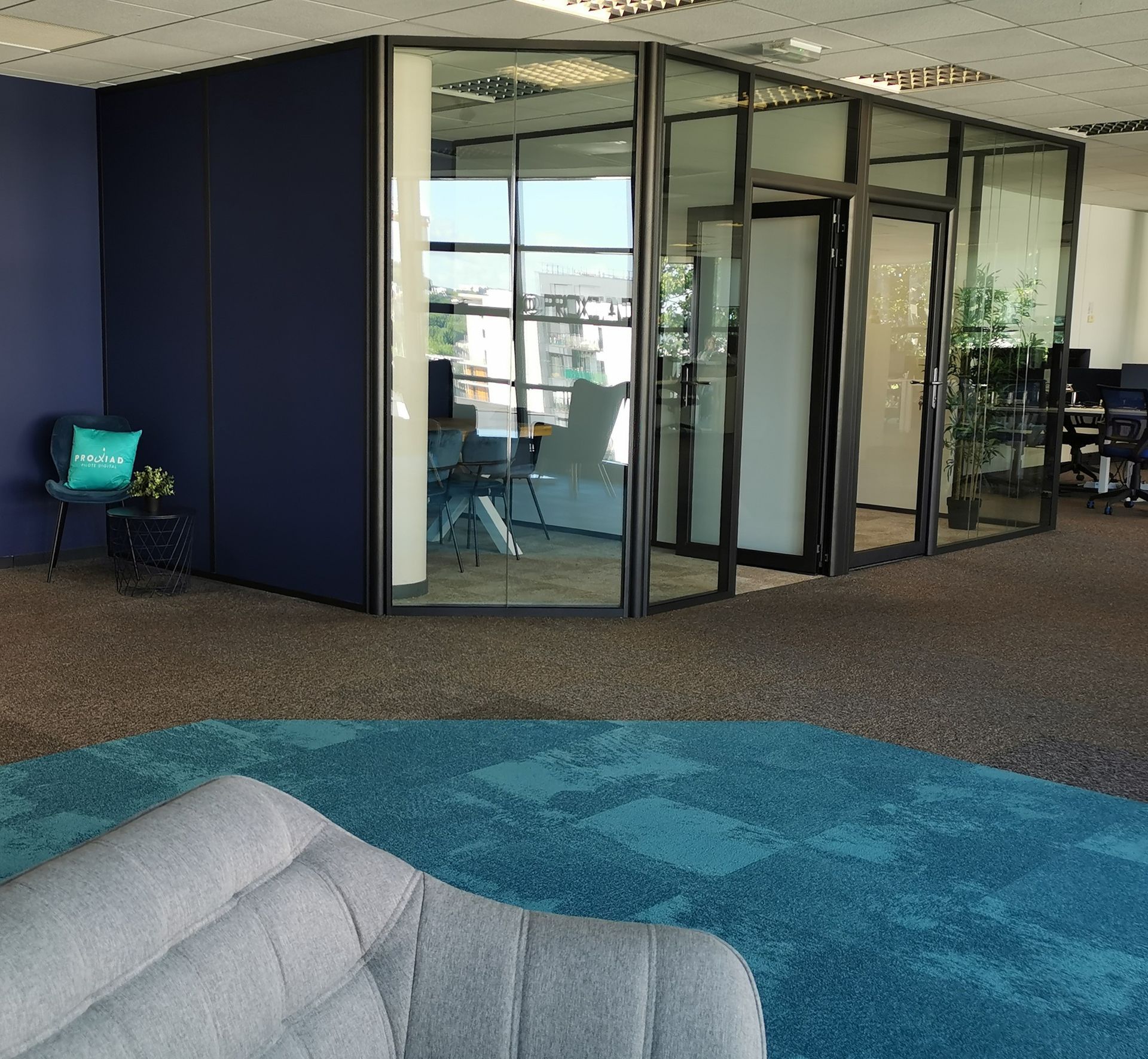 Intérieur de bureau avec bureau aux parois vitrées, tapis bleu, une chaise et un coin salon avec une chaise grise.