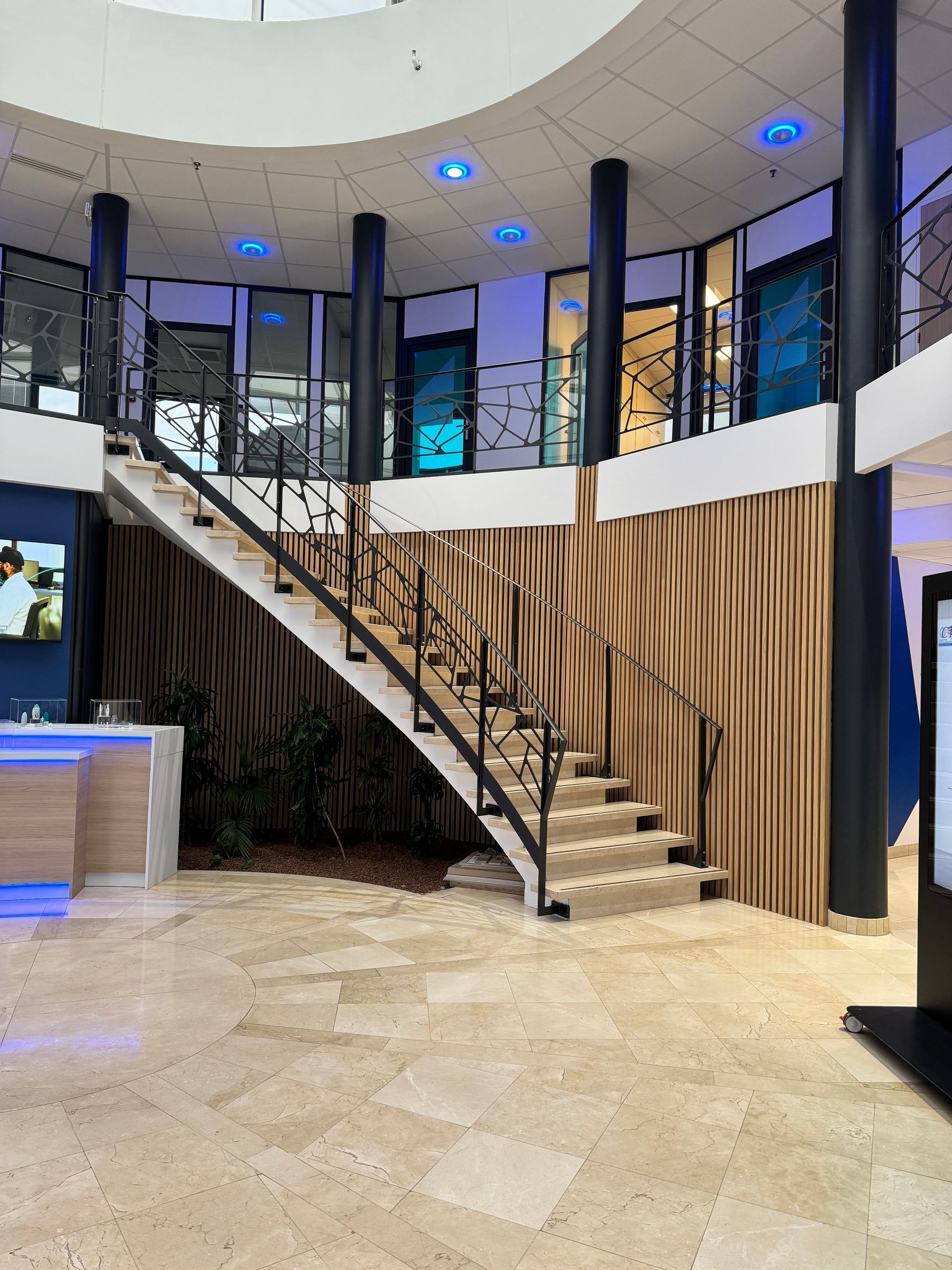 Escalier dans un bureau moderne, sols en pierre beige, rampes noires et boiseries au mur.