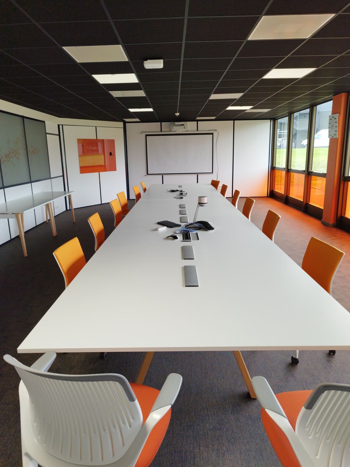 Salle de conférences avec une longue table blanche, des chaises orange et un tableau blanc. Plafond noir et mur orange.