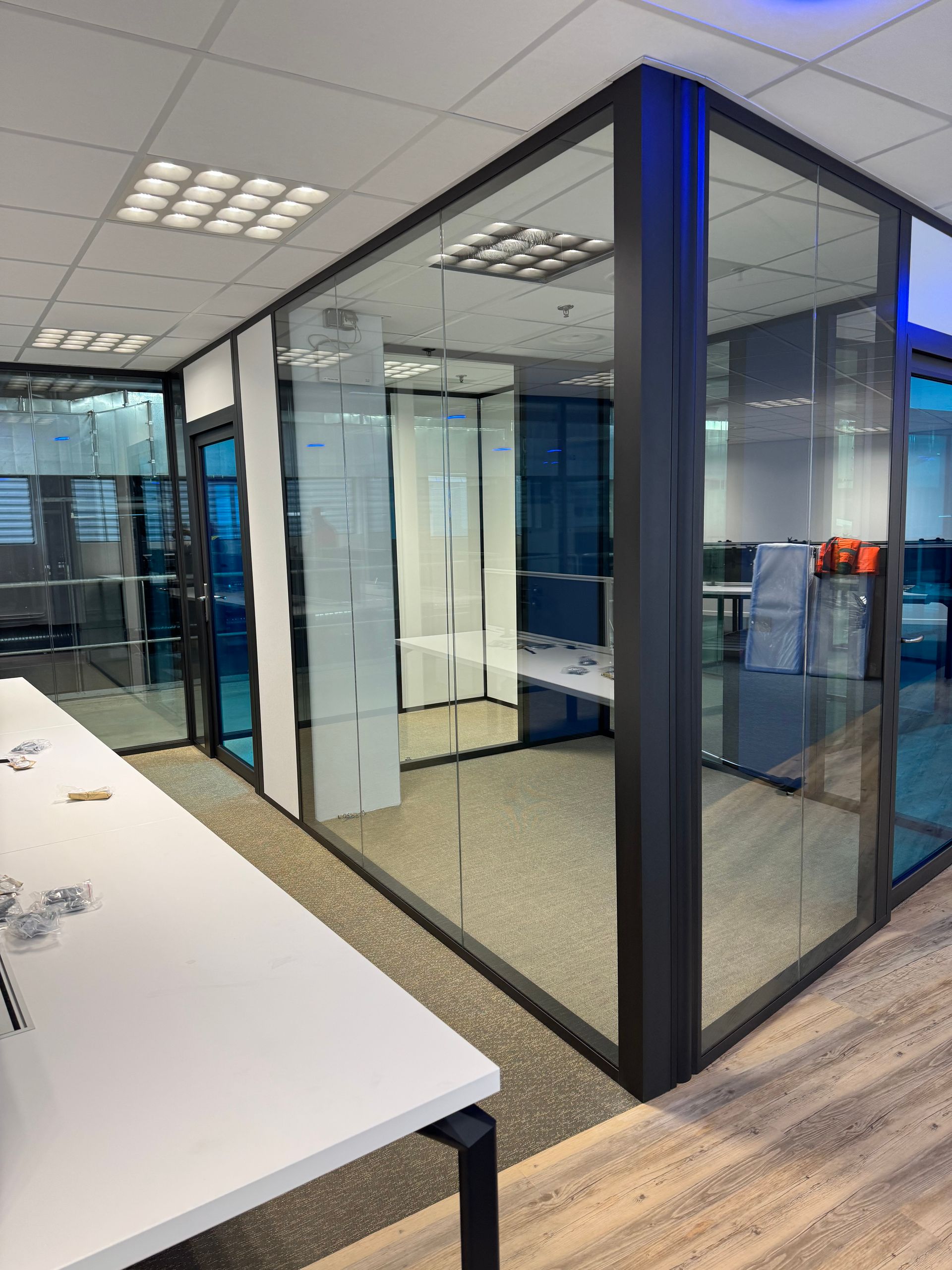 Bureau vitré avec un bureau blanc et une petite salle de conférences en arrière-plan. Éclairage fluorescent.