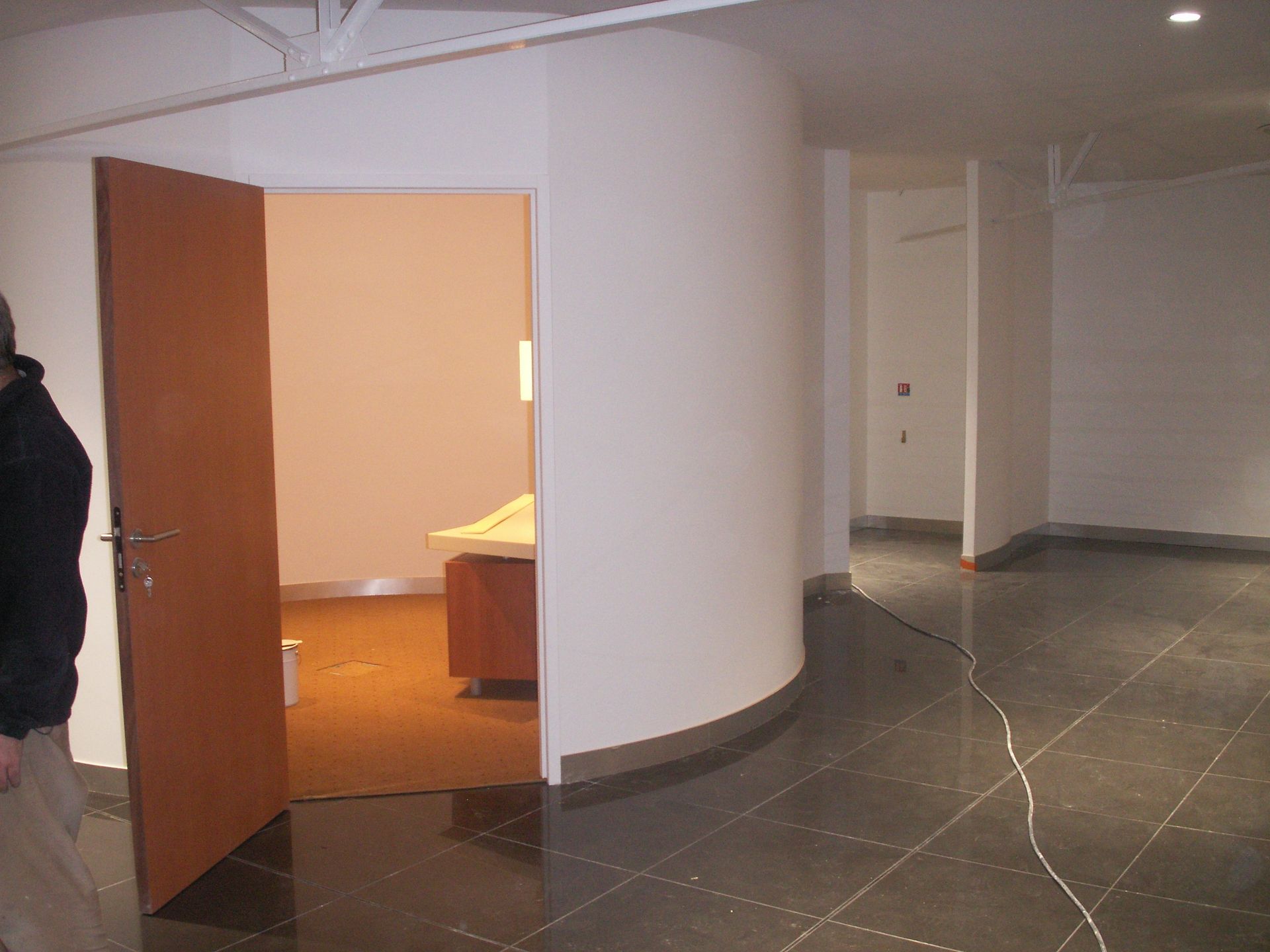 Une personne se tient près d'une porte ouverte donnant sur une pièce. Mur blanc incurvé, sol sombre.