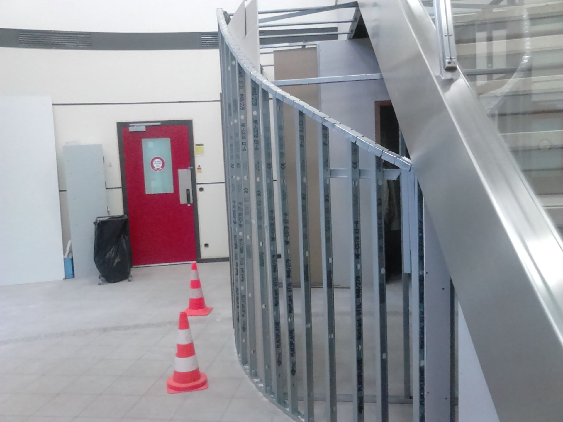 Escalier métallique incurvé avec rampe. Porte rouge, cônes de signalisation orange et décor de chantier.