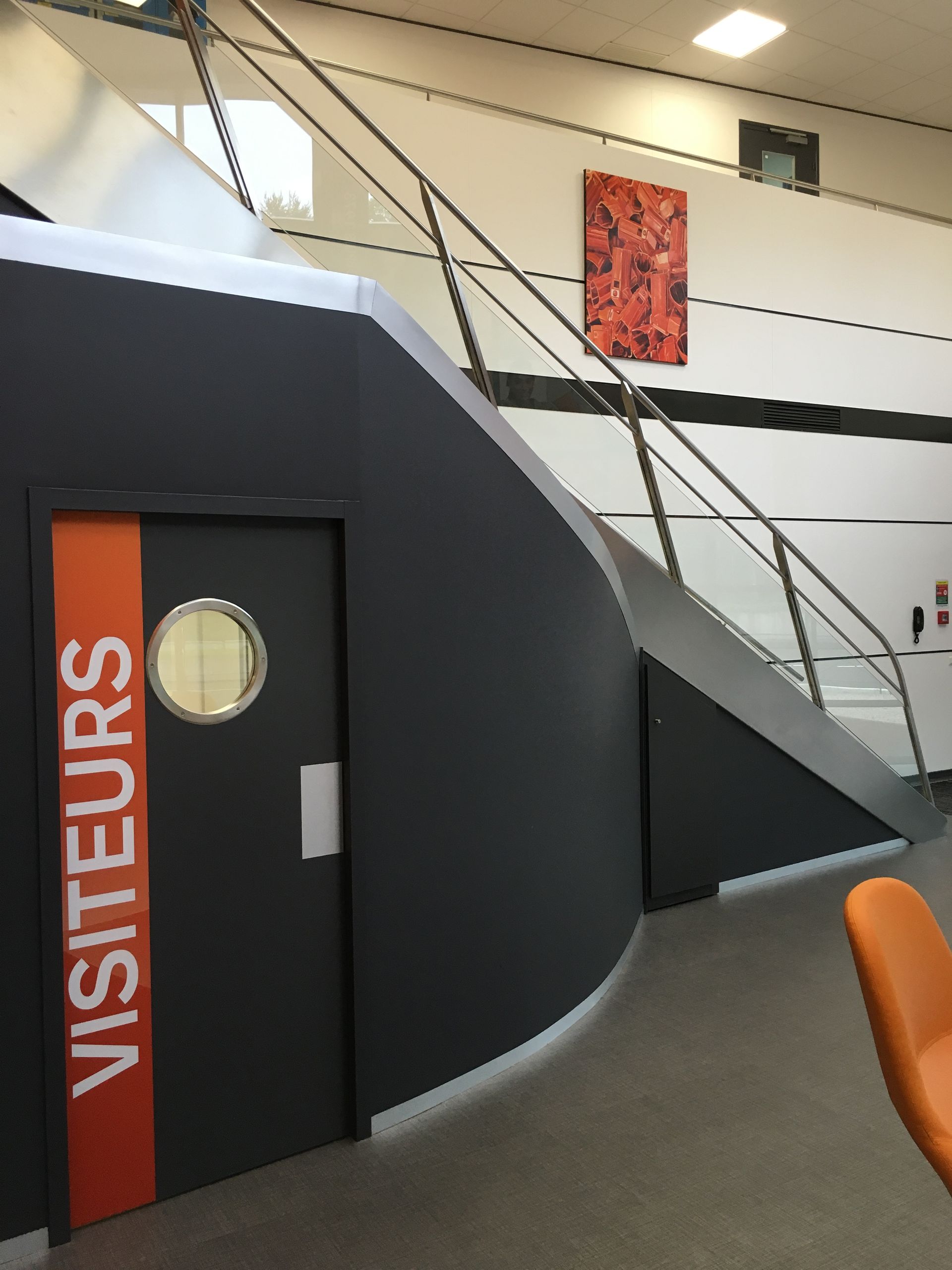 Mur incurvé gris foncé avec l'inscription « VISITEURS » sur une bande orange et une porte hublot ; escalier avec une rampe en métal.