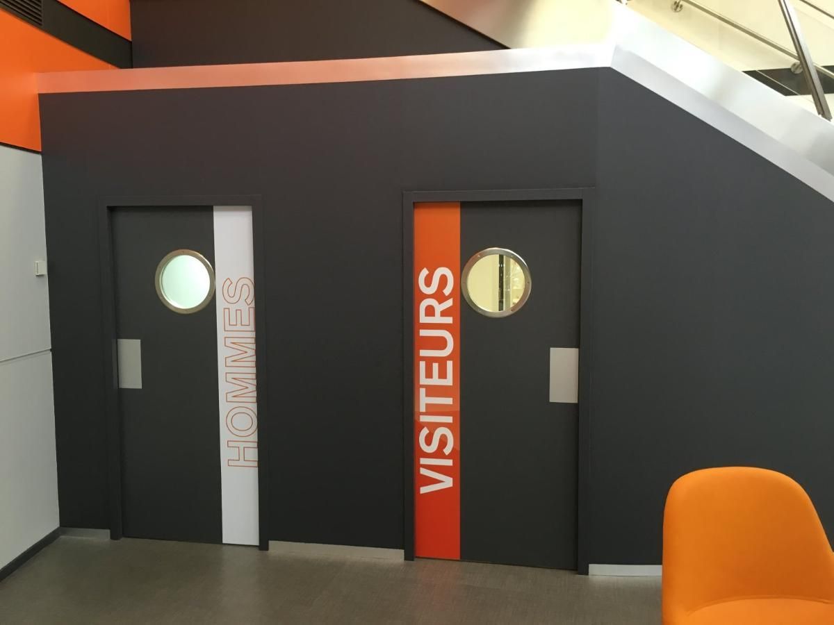 Deux portes gris foncé étiquetées VISITEURS et SALLES avec des fenêtres circulaires, dans un bâtiment aux accents orange.