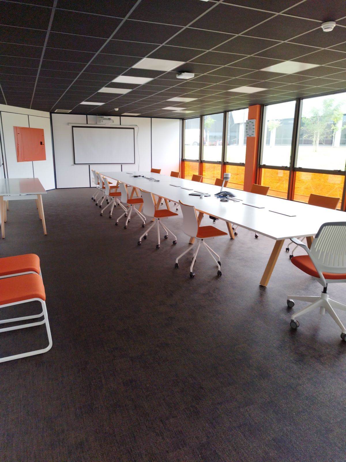 Salle de conférence avec longue table blanche, chaises orange, écran de projection, grandes fenêtres et sol foncé.