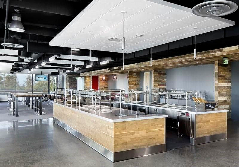 Cafétéria moderne avec postes de restauration, comptoirs en bois clair et en acier inoxydable et sol en béton gris.