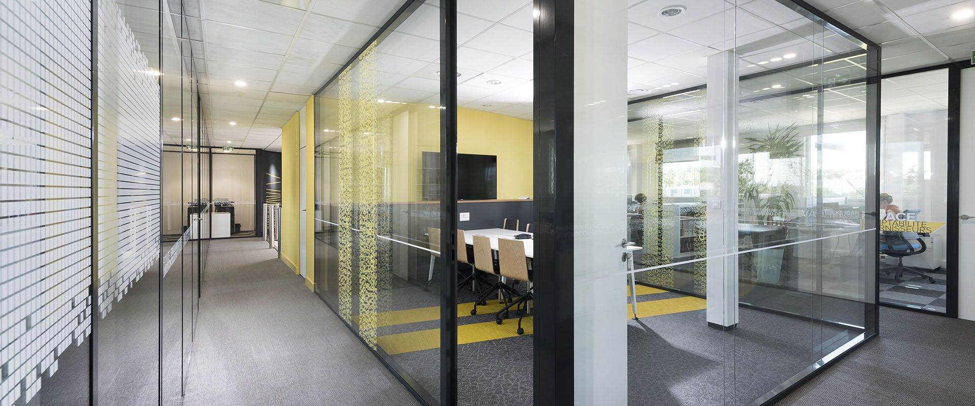 Couloir de bureau avec murs et portes en verre, montrant les salles de réunion et les bureaux, couleurs neutres.