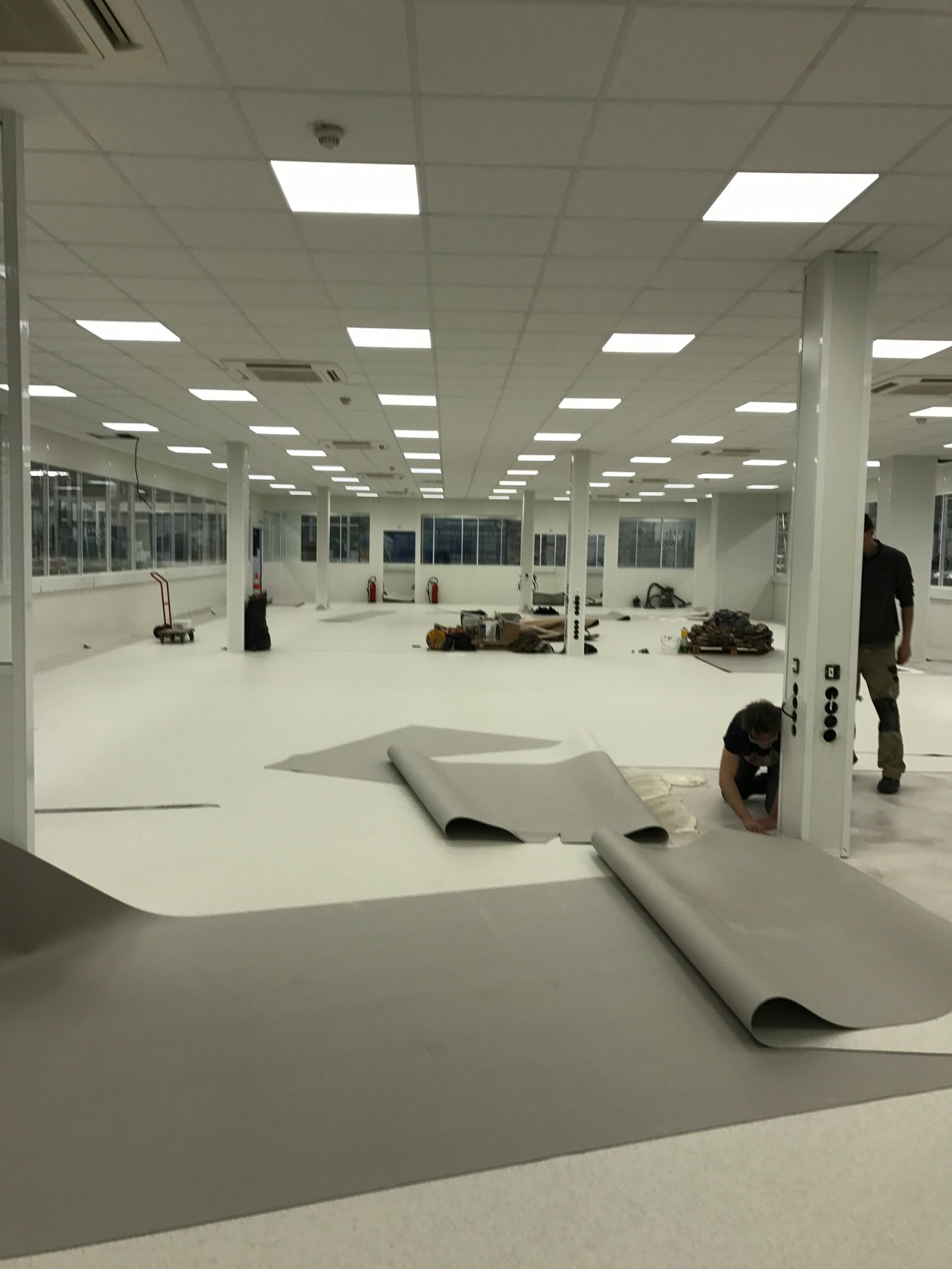 Une grande pièce lumineuse avec un sol gris en cours d'installation. Des ouvriers sont présents.