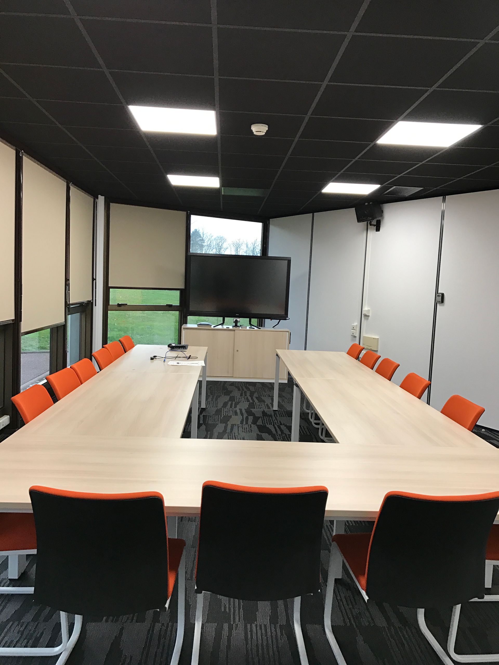 Salle de conférence avec une table en forme de U, des chaises orange et un grand écran.