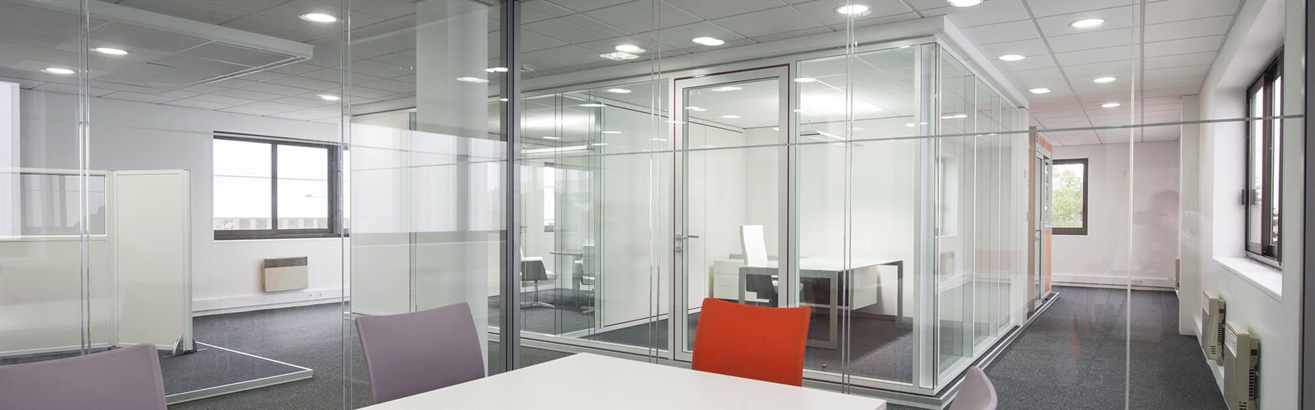 Intérieur d'un bureau moderne avec parois vitrées, murs blancs et moquette grise. Touches orange et violettes.