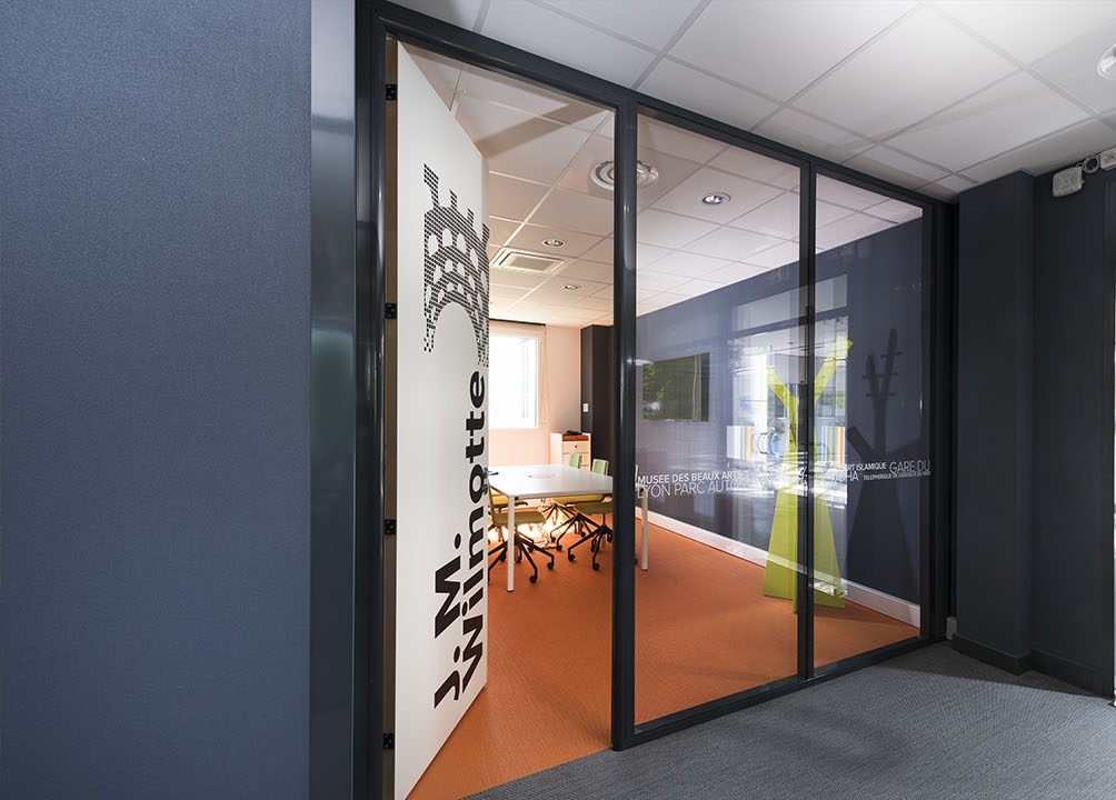 Salle de conférences vitrée avec porte ouverte, sol orange et table blanche. Couleurs : bleu, gris et blanc.