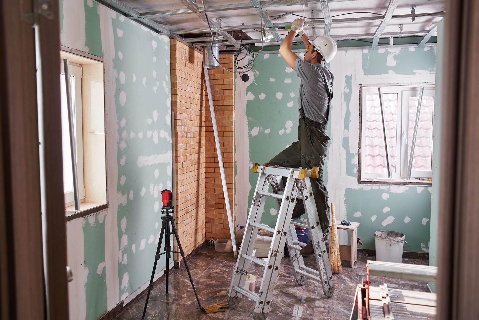 Un ouvrier, juché sur une échelle, installe des plaques de plâtre dans une pièce en rénovation.