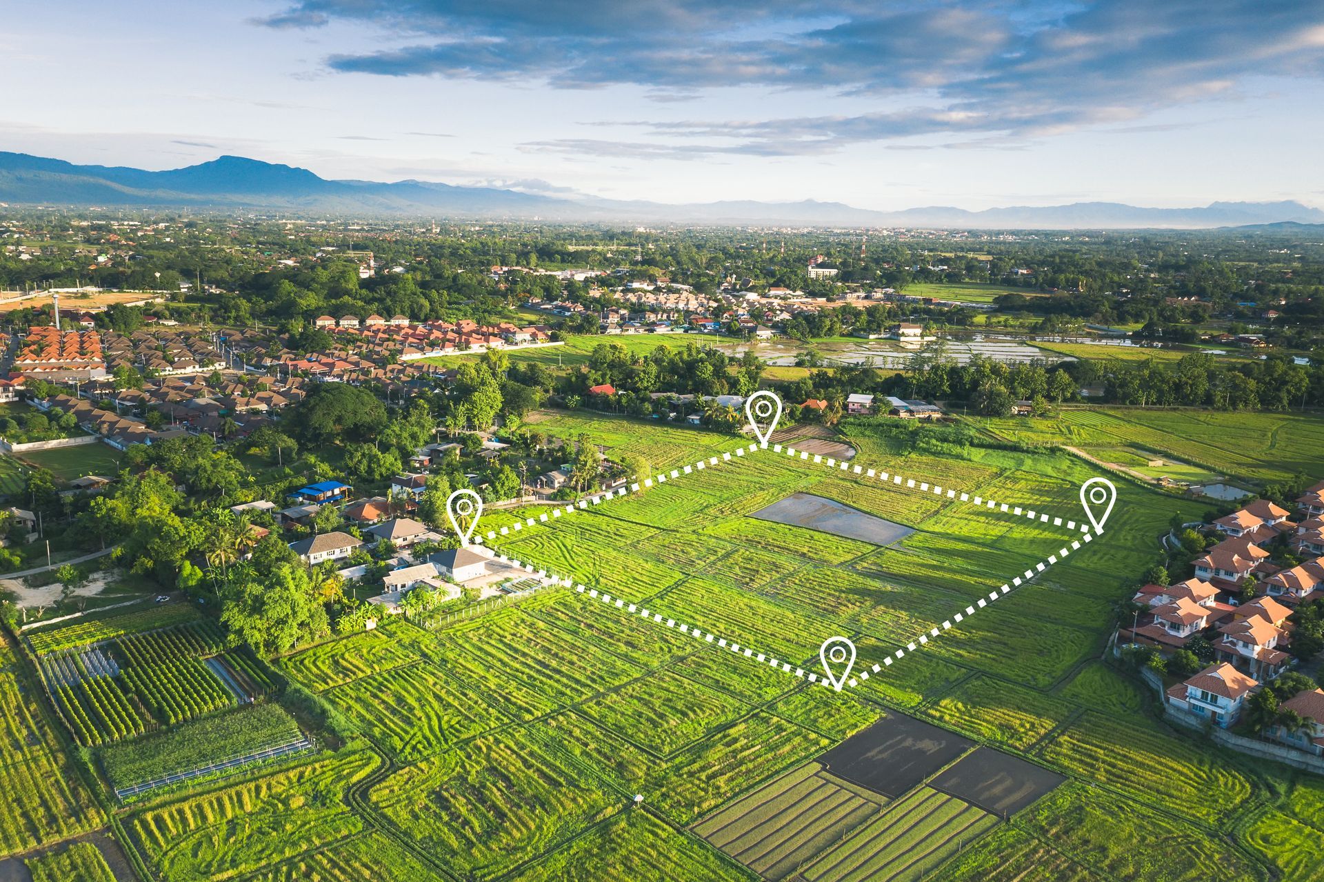 Vue aérienne d'un champ agricole verdoyant entouré de maisons et d'un paysage urbain sous un ciel lumineux.