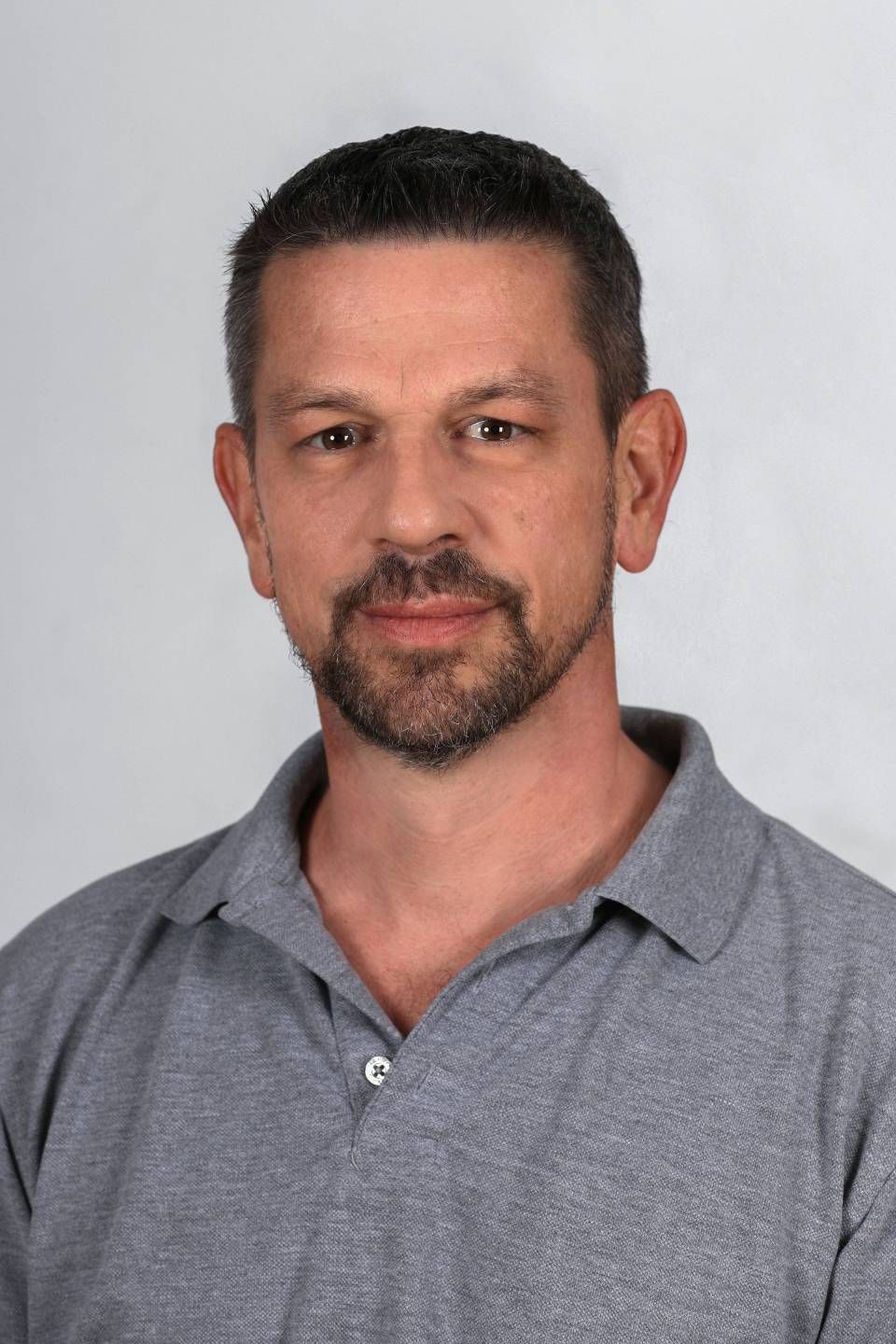 Eine Person mit kurzen, dunklen Haaren und einem Spitzbart, die ein graues Poloshirt trägt, vor einem schlichten, hellen Hintergrund.