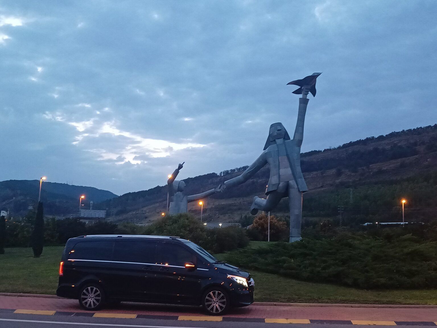 Une Mercedes noire garée en bord de route au crépuscule, avec une grande sculpture métallique et des collines en arrière-plan.