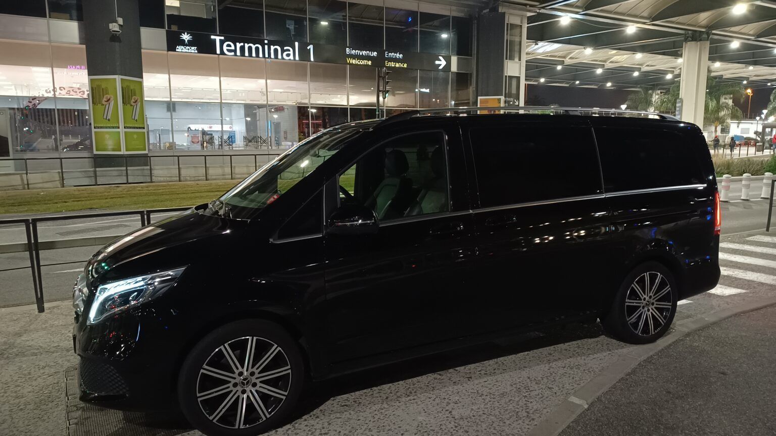 Une Mercedes noire garée devant un terminal la nuit, sous des lumières vives