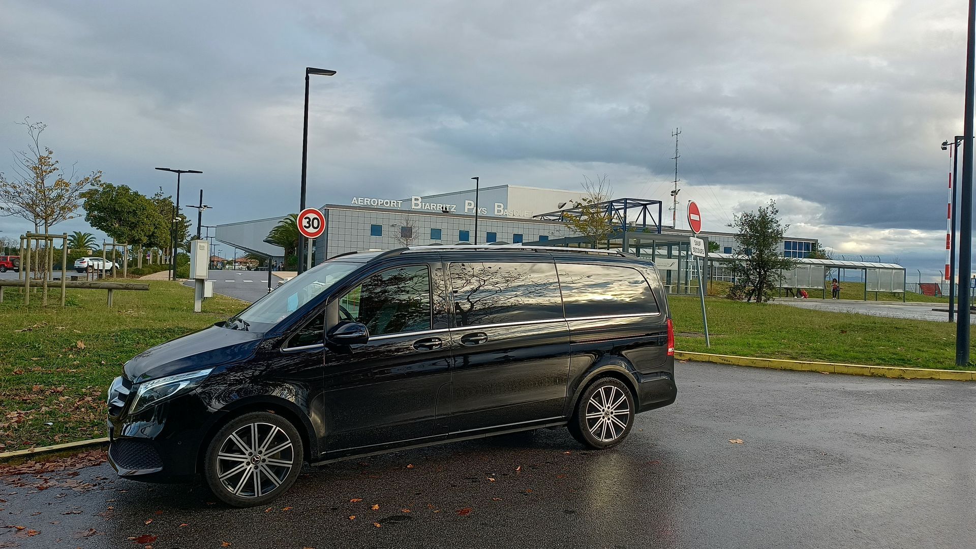 Une Mercedes noire est garée sur un terrain gravillonné devant un grand bâtiment historique en bord de mer.