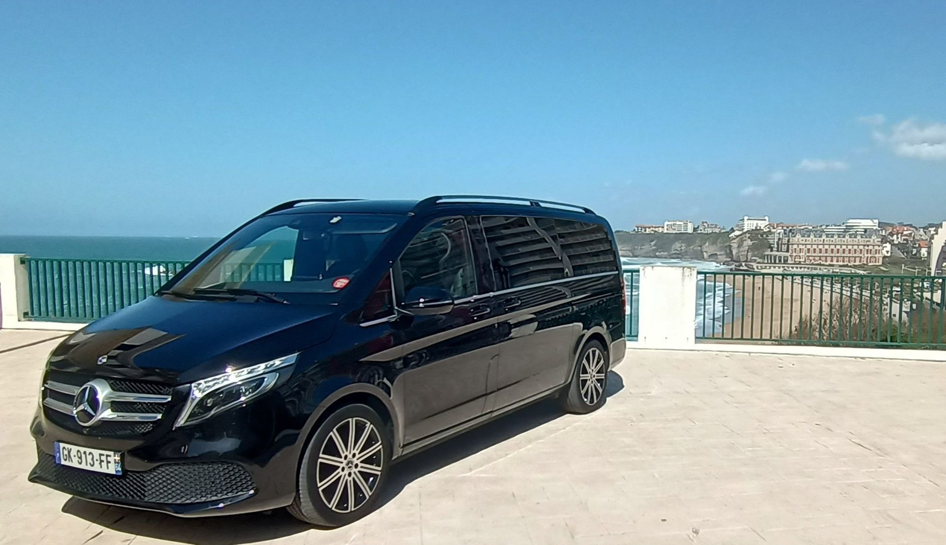 Une Mercedes noire est garée sur un belvédère ensoleillé au bord de l'eau, avec des immeubles en arrière-plan.