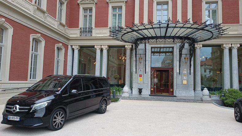 Une Mercedes noire est garée sous l'entrée d'un grand hôtel aux murs de briques rouges et avec une verrière.