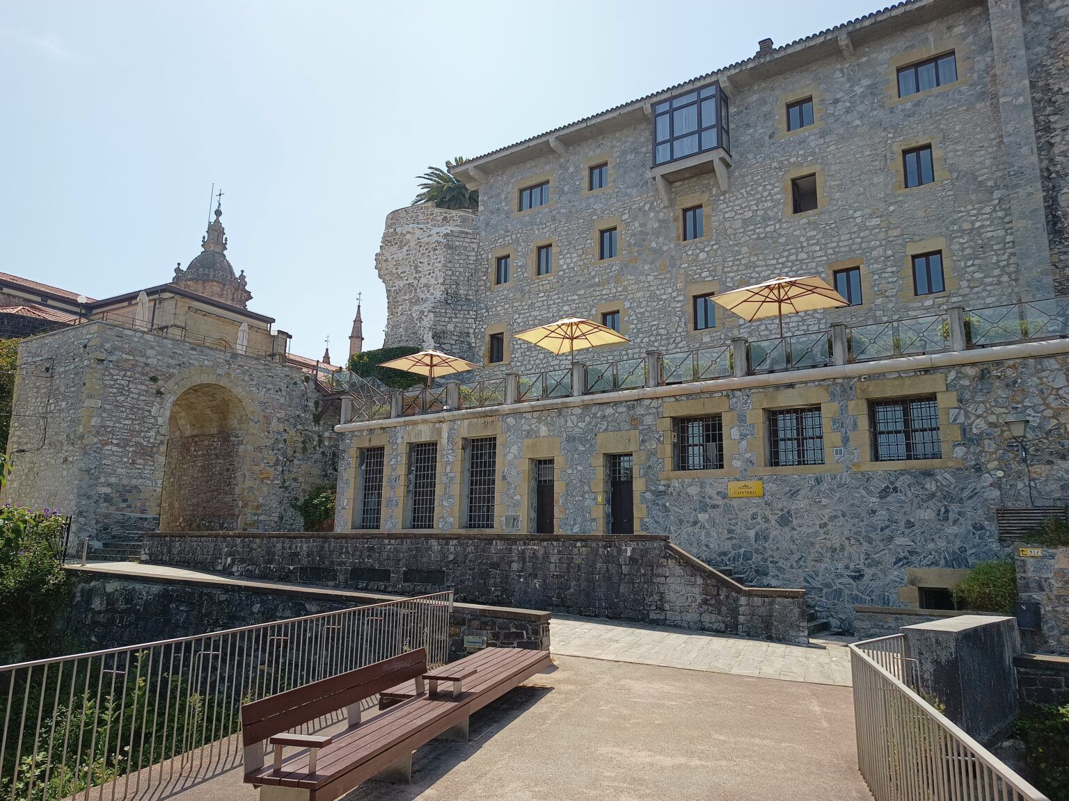 Cour en pierre avec bancs et parasols à côté d'un bâtiment historique ressemblant à un château, sous un ciel bleu limpide