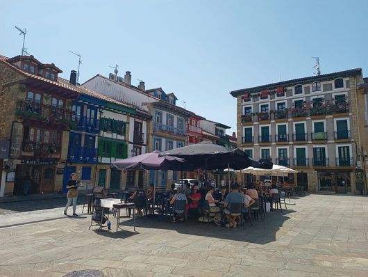 Terrasses de café sous des parasols sur une place ensoleillée entourée de bâtiments colorés.