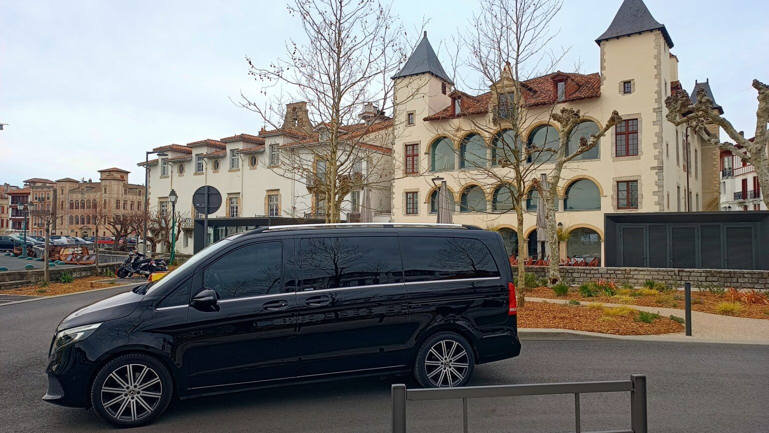 Une Mercedes noire est garée dans une rue de la ville, devant un grand immeuble couleur crème avec des tours et des arbres dénudés.
