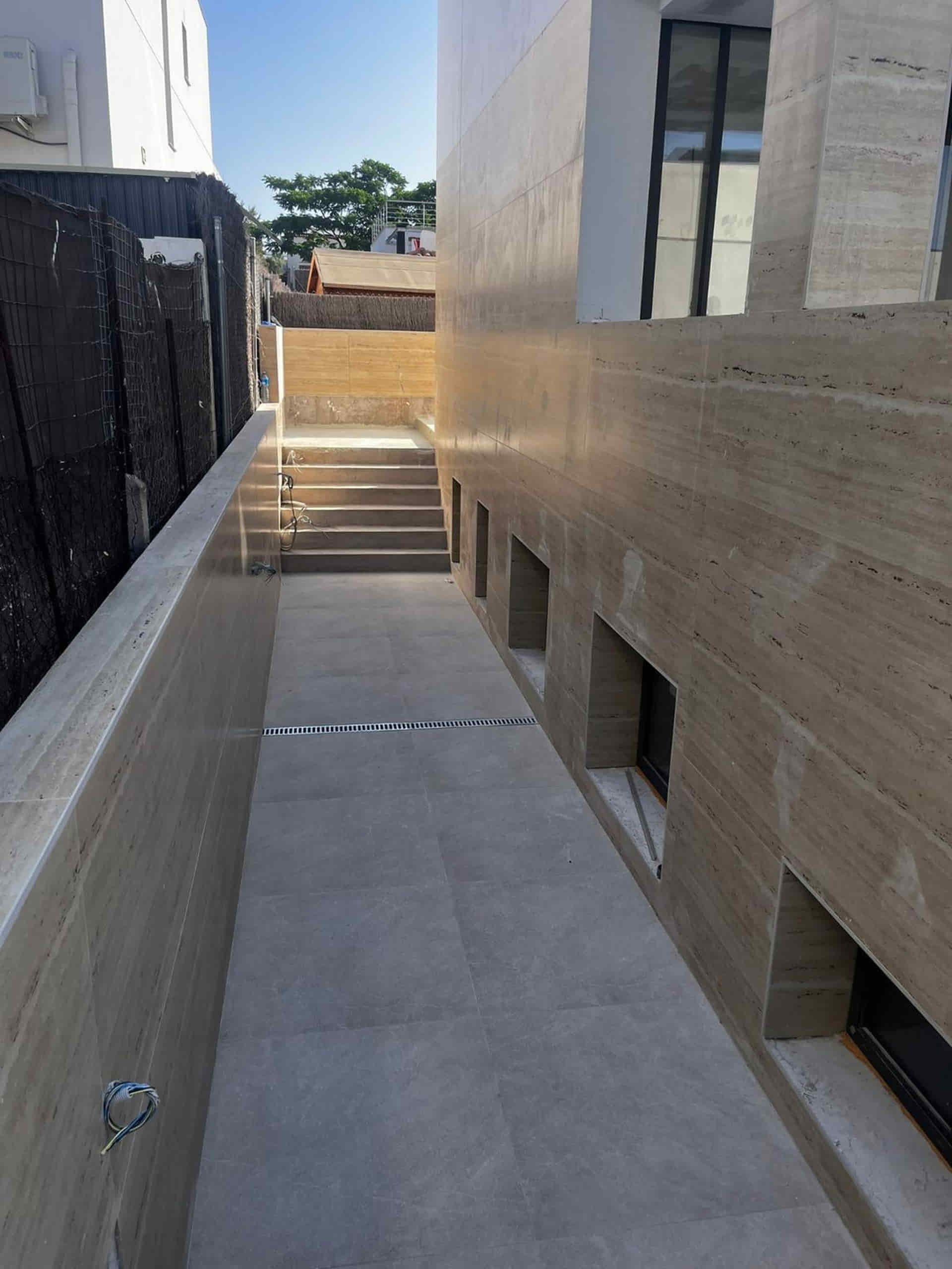 Callejón estrecho con escaleras. Muros de piedra, ventanas a la derecha, barandilla a la izquierda. Luz solar intensa.