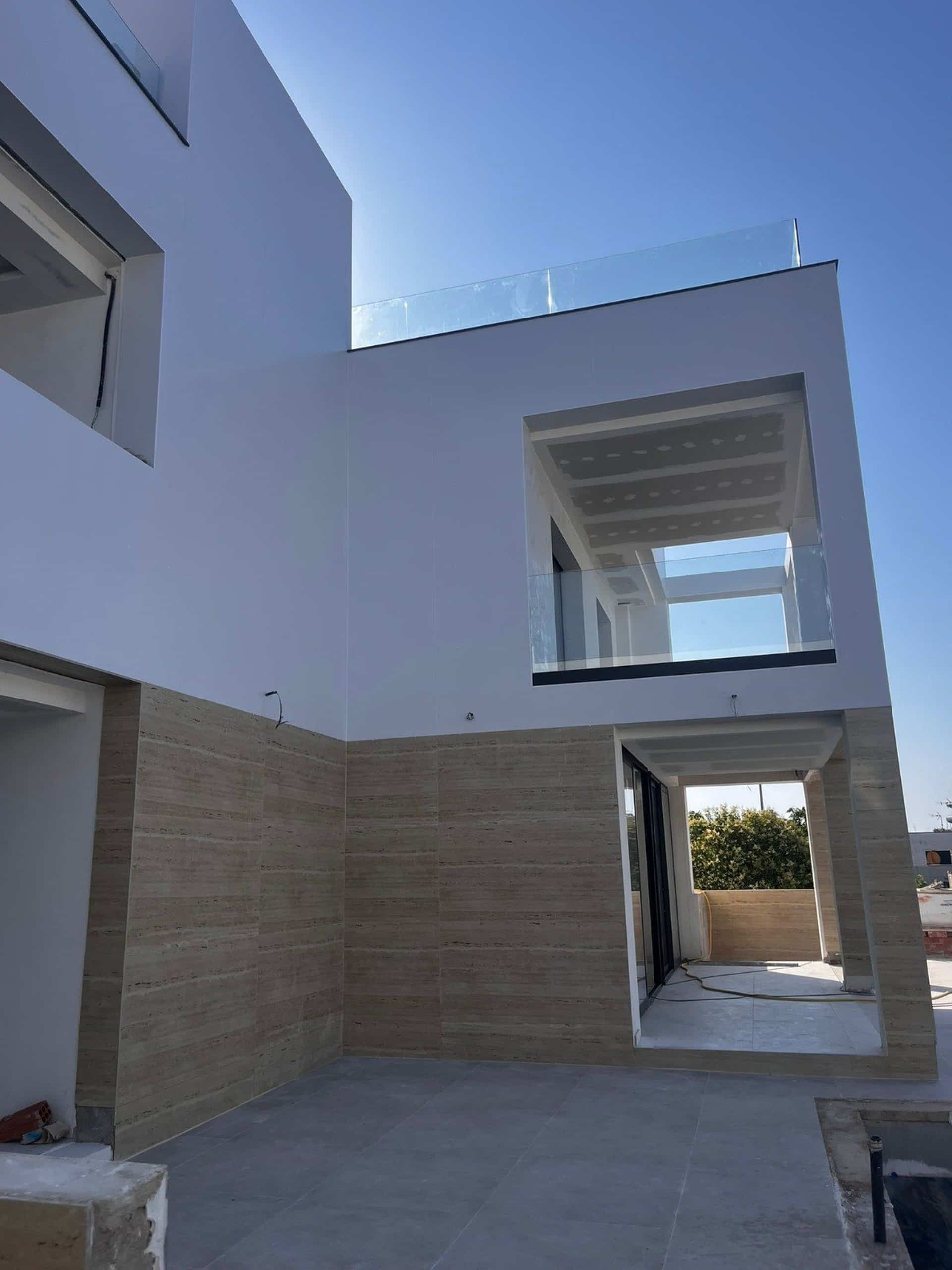 Casa moderna de color blanco y tostado con barandillas de cristal y balcón abierto bajo un cielo azul claro.