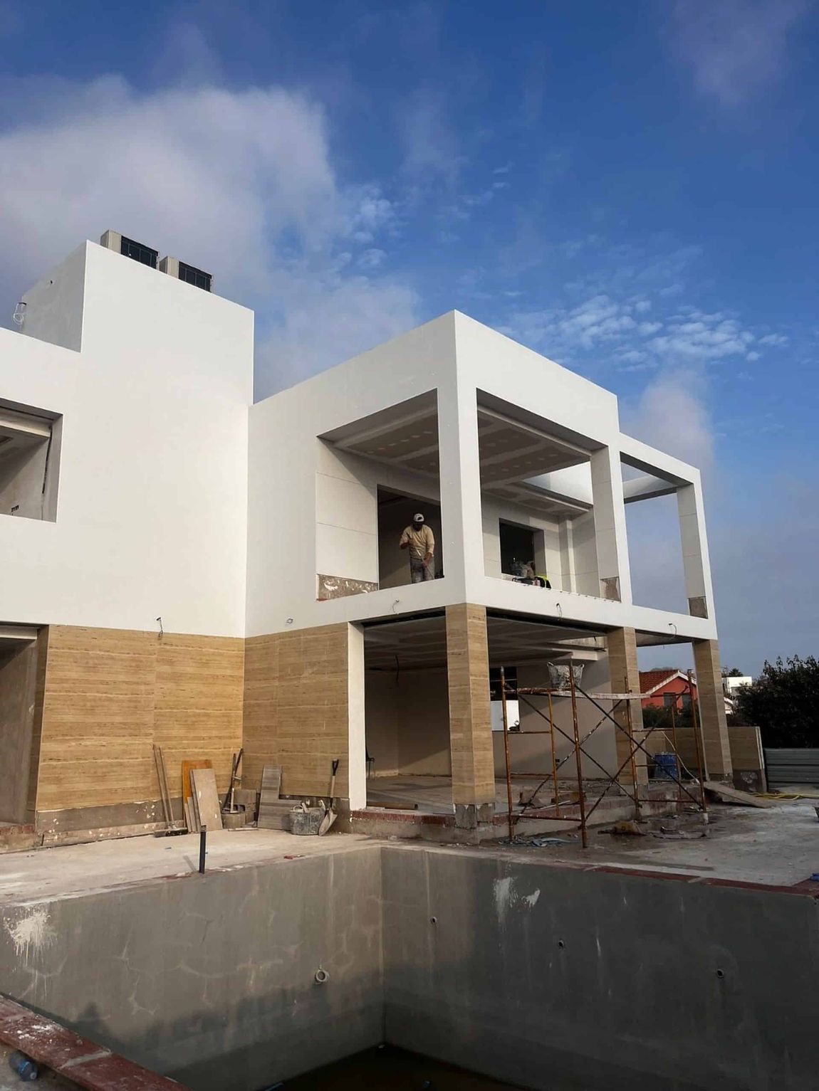 Edificio de dos pisos en construcción con una persona parada en una puerta; una piscina en primer plano, cielo azul.