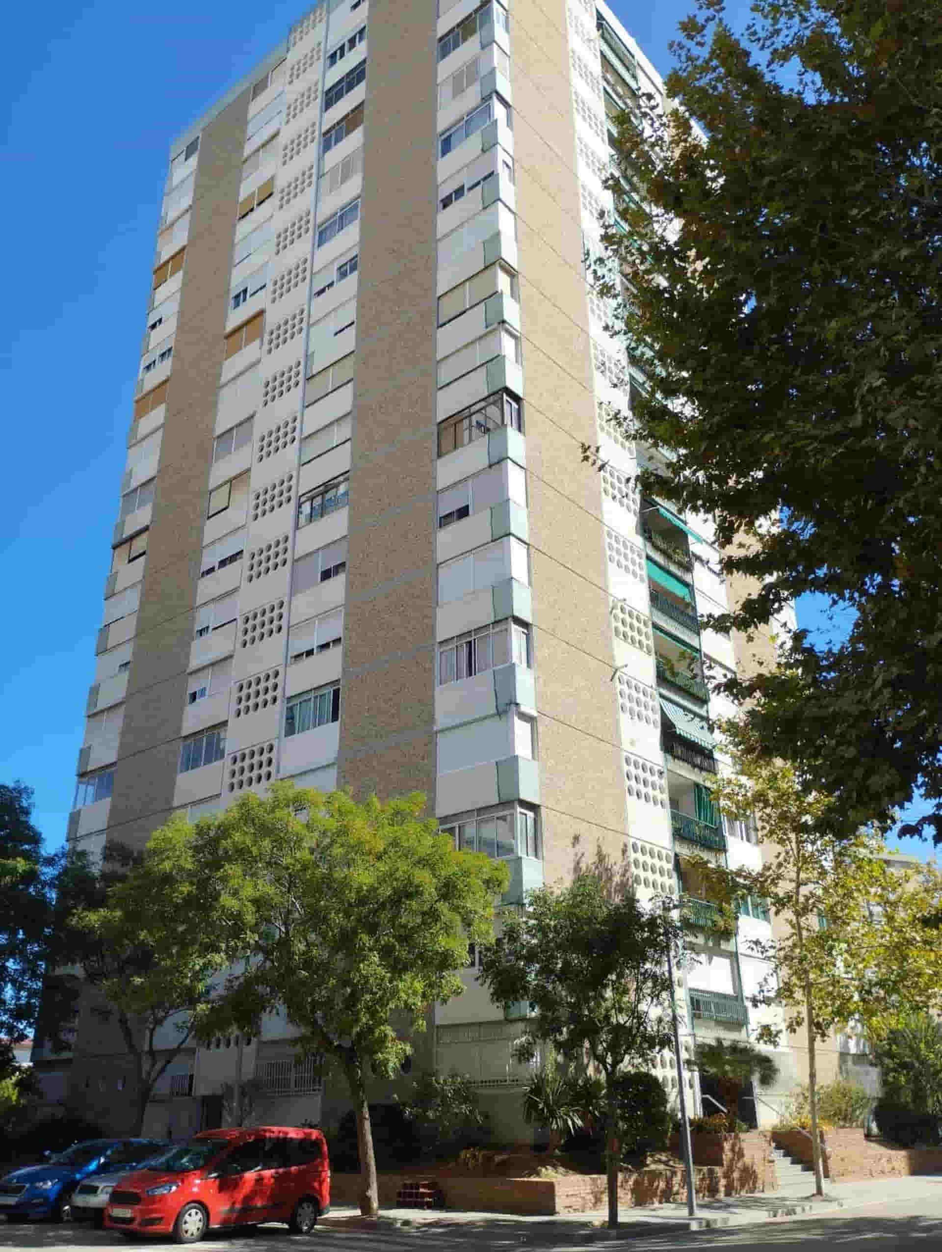 Edificio alto de apartamentos de color beige y blanco, con muchas ventanas, coches aparcados en el frente.