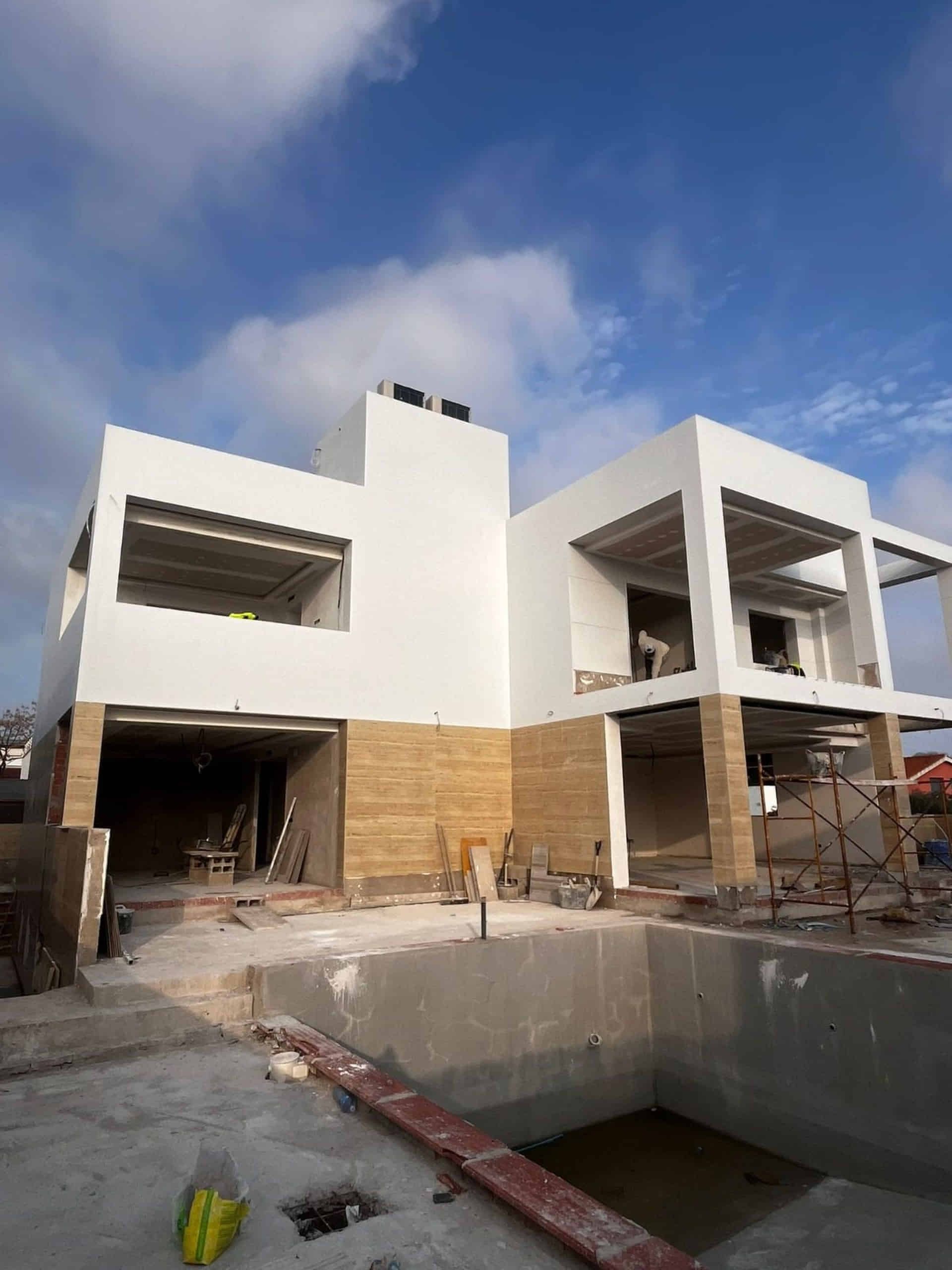 Construcción de una casa moderna de color blanco con un nivel inferior de piedra beige y una piscina, bajo un cielo azul.