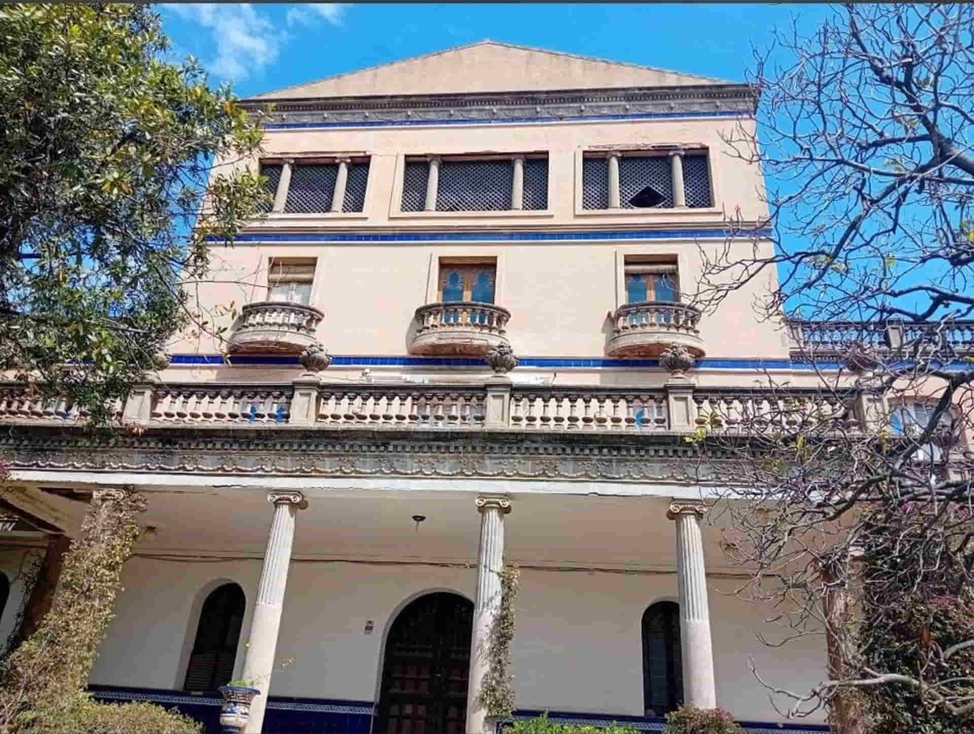 Mansión de estuco blanco y azul con columnas, balcones y tres pisos, bajo un cielo azul.