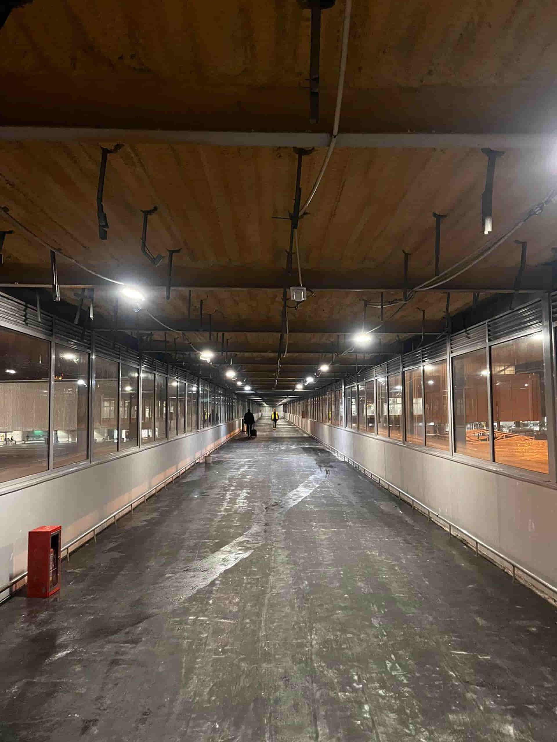 Largo pasillo interior con paneles de vidrio, luces de techo y piso oscuro.