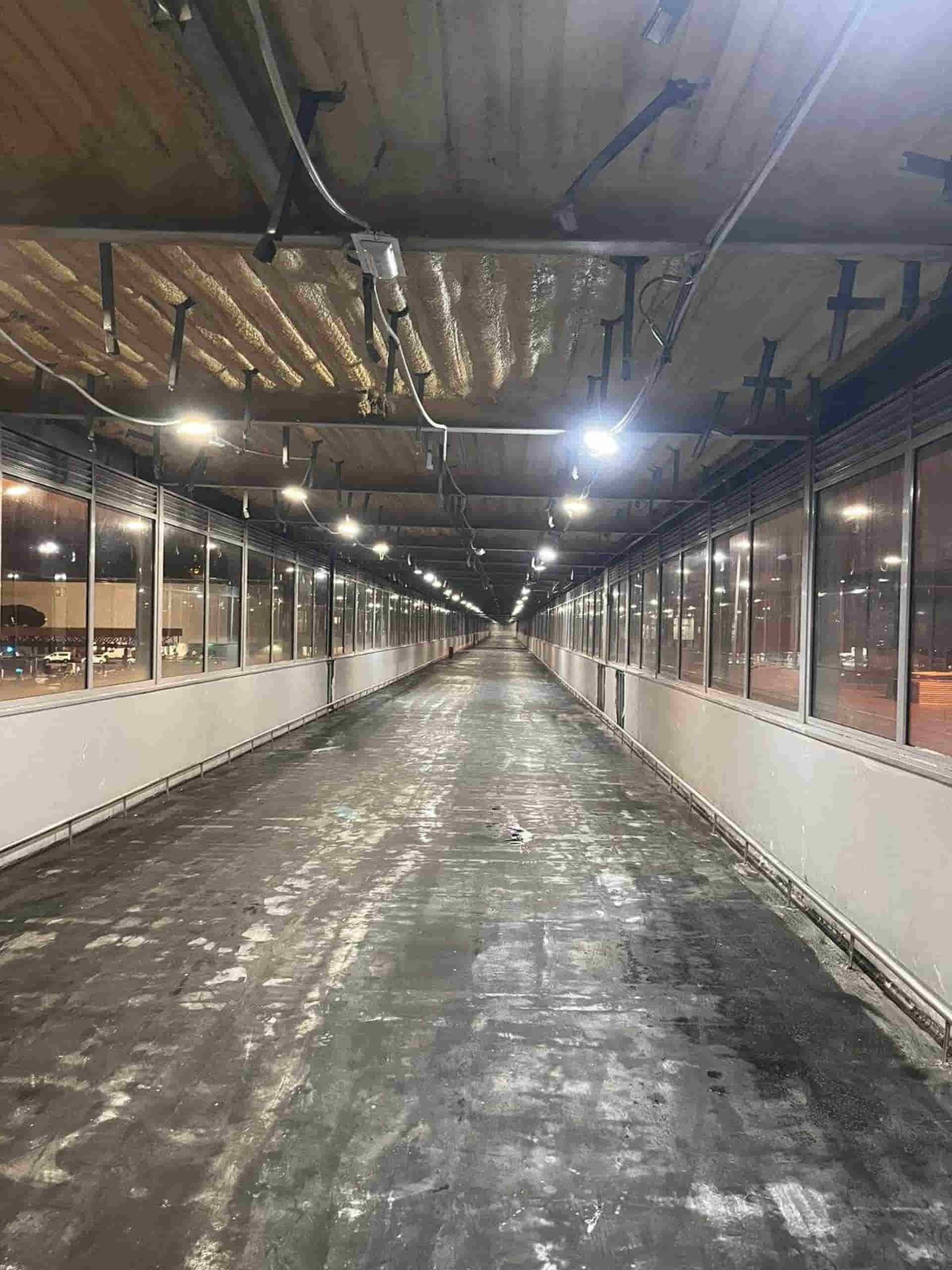 Largo paseo peatonal vacío, iluminado por luces cenitales y con ventanas a ambos lados.