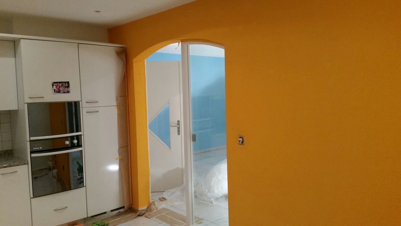 Gelb gestrichene Wand mit gewölbtem Türbogen, weissen Schränken und einer Glastür, die in einen hellblauen Raum führt.