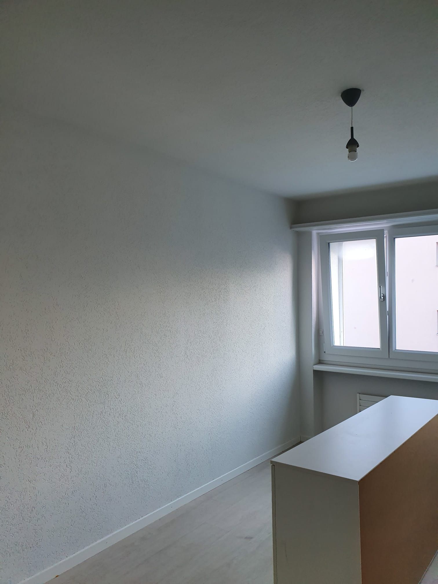 Leerer Raum mit weiss strukturierten Wänden, Fenster, weissem Schrank, Lampe und Holzboden.