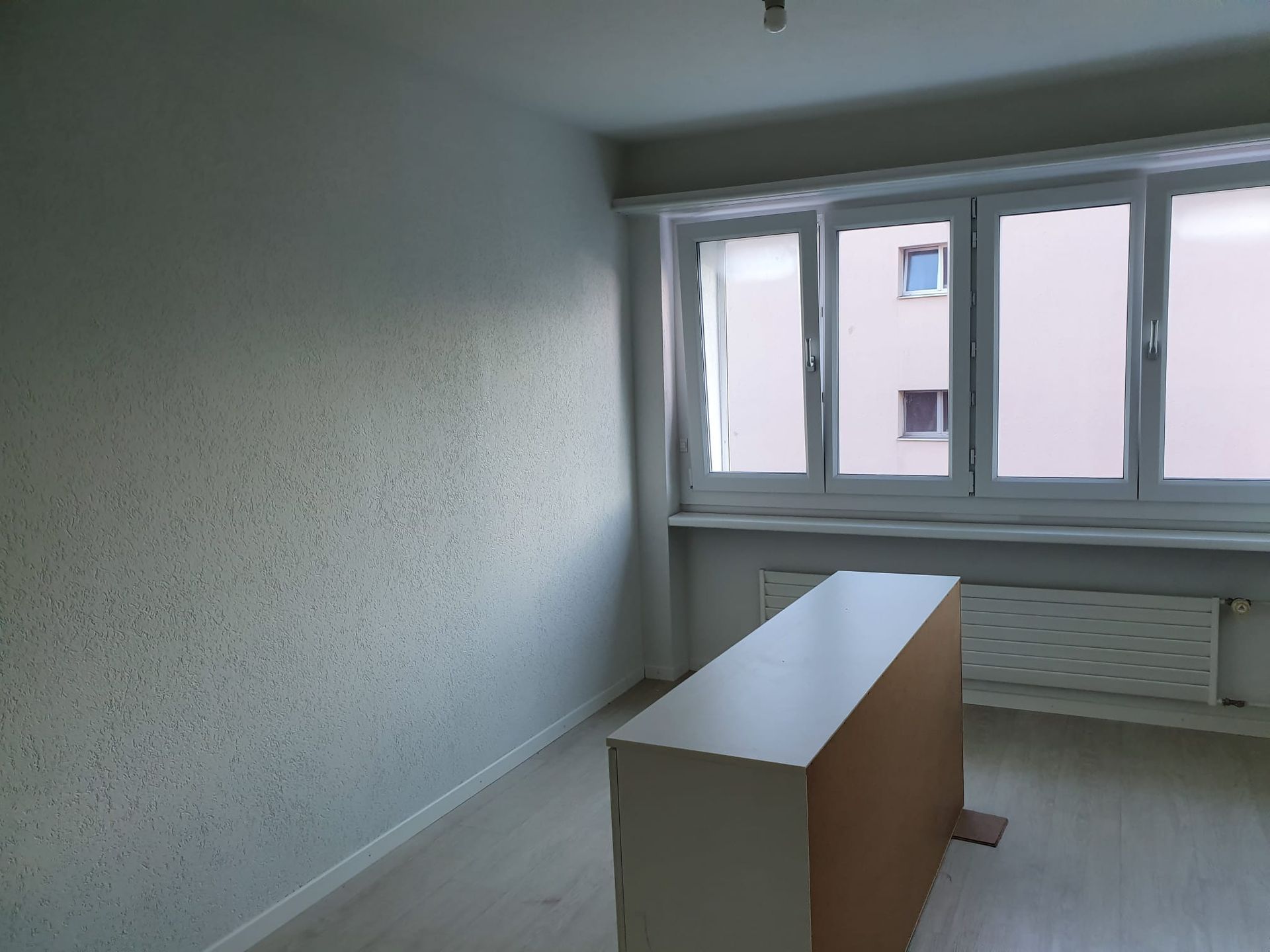 Leeres Zimmer mit grossem Fenster, weisser Kommode und Heizkörper.