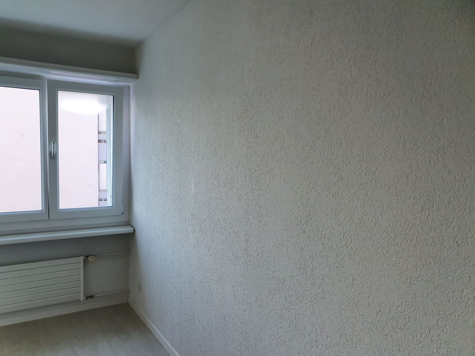Leeres Zimmer mit strukturierten weissen Wänden, einem Fenster und einem Heizkörper. Holzboden.