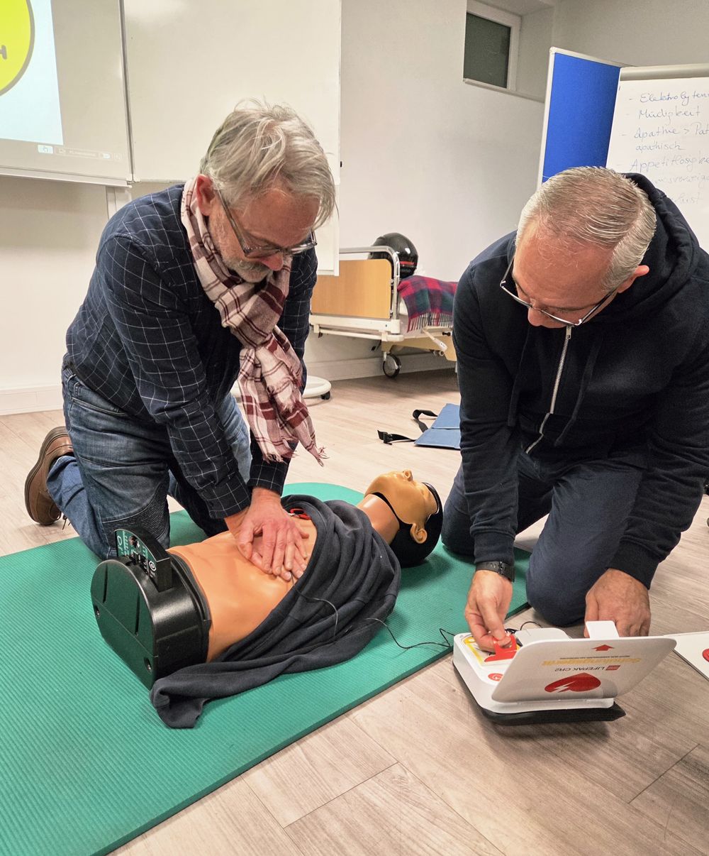 Zwei Personen üben die Herz-Lungen-Wiederbelebung an einer Übungspuppe, eine davon benutzt einen AED. Übungsumgebung: Innenräume.