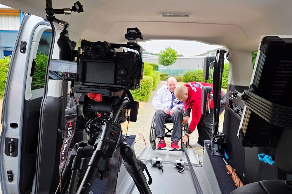 Camera filming two people in wheelchairs inside a van, with one person in red helping.