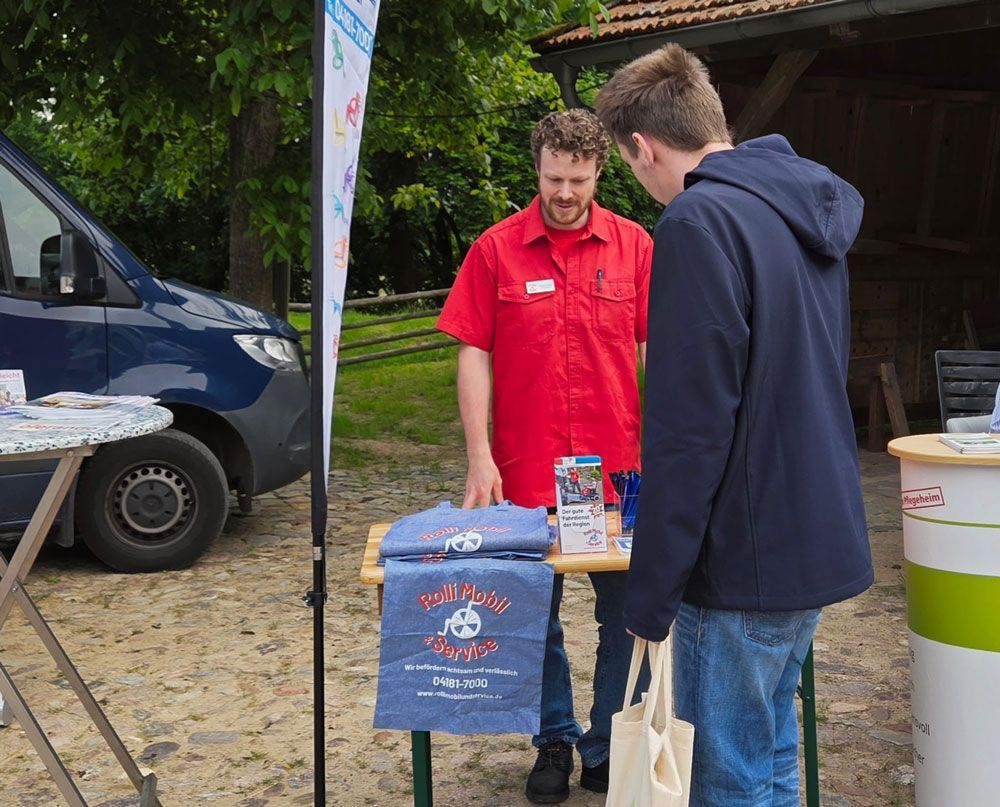 Two people at an outdoor booth, one in a red shirt behind a table, beside a blue van and a tote bag