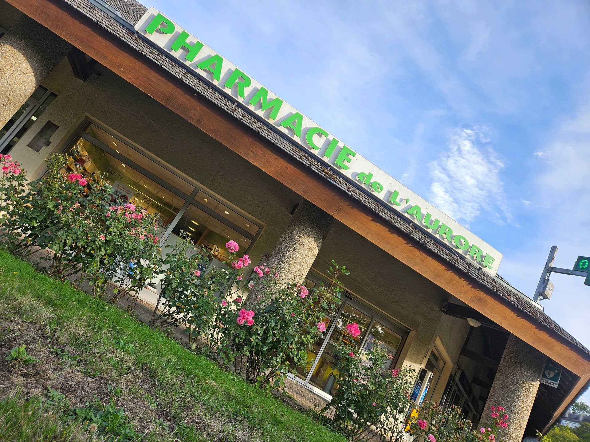 Vue inclinée de la façade d'une pharmacie avec une enseigne verte, des colonnes en pierre, des fleurs roses et un ciel bleu.