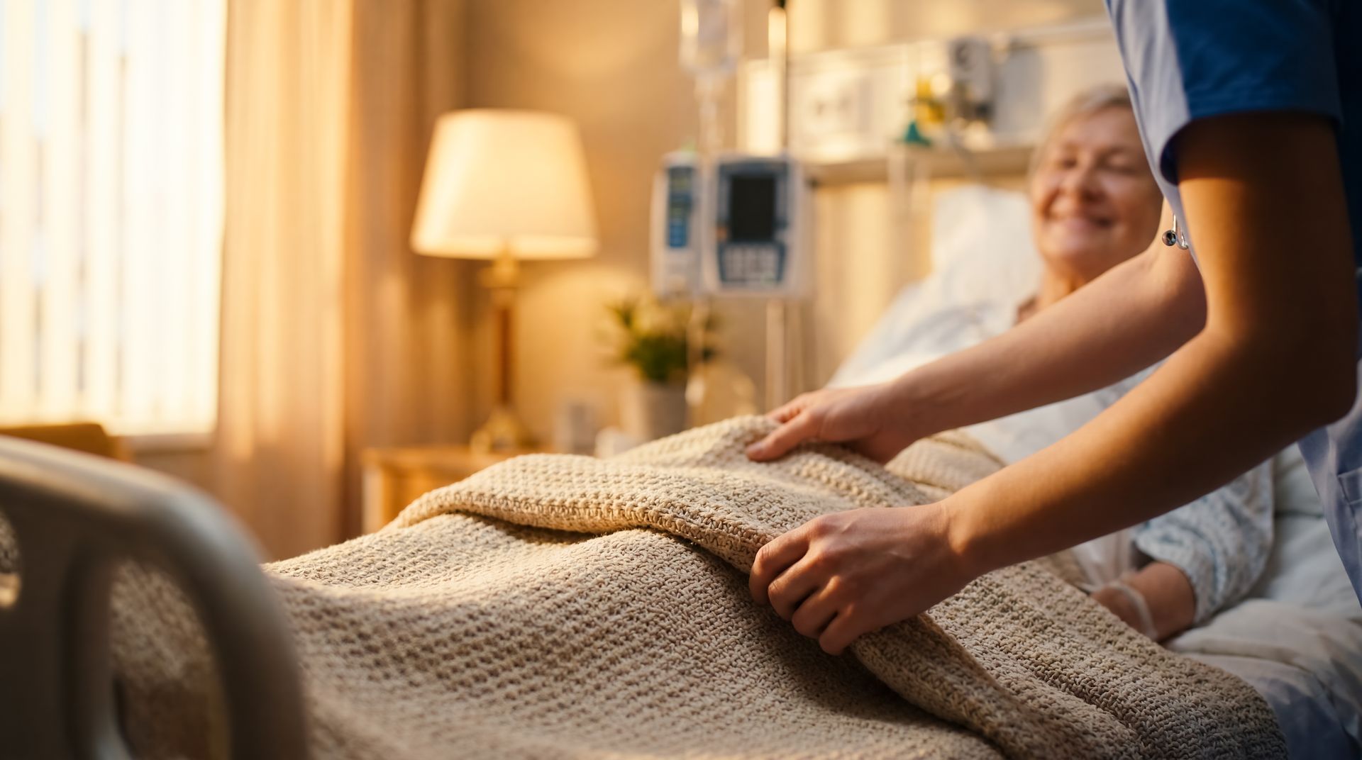 Une soignante borde un patient souriant d'une couverture dans une chambre chaleureuse et doucement éclairée