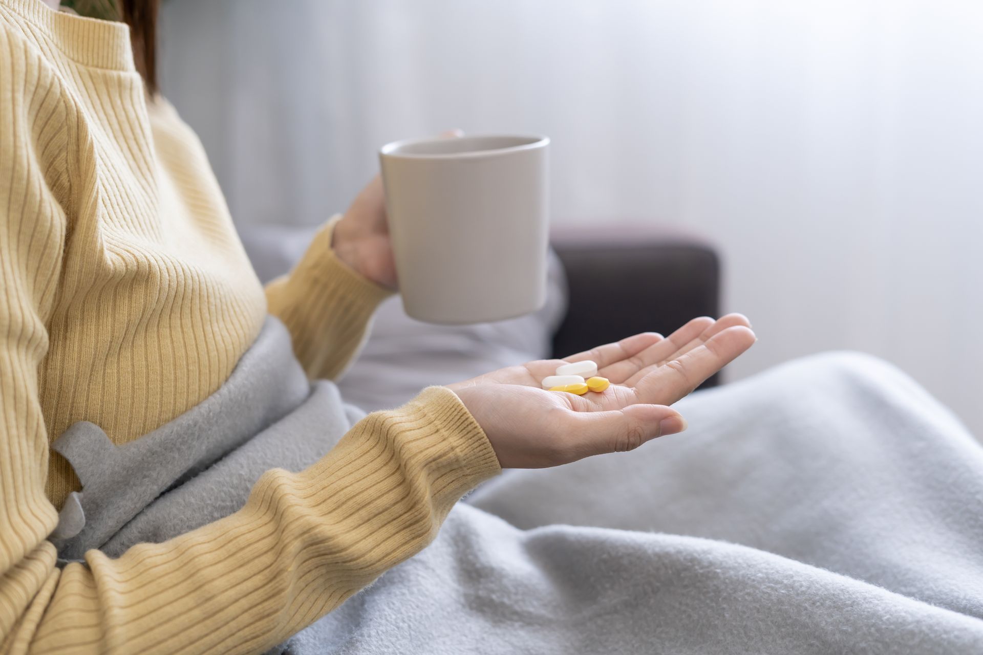 Une personne en pull jaune, tenant une tasse et des pilules, est assise sous une couverture grise sur un canapé.