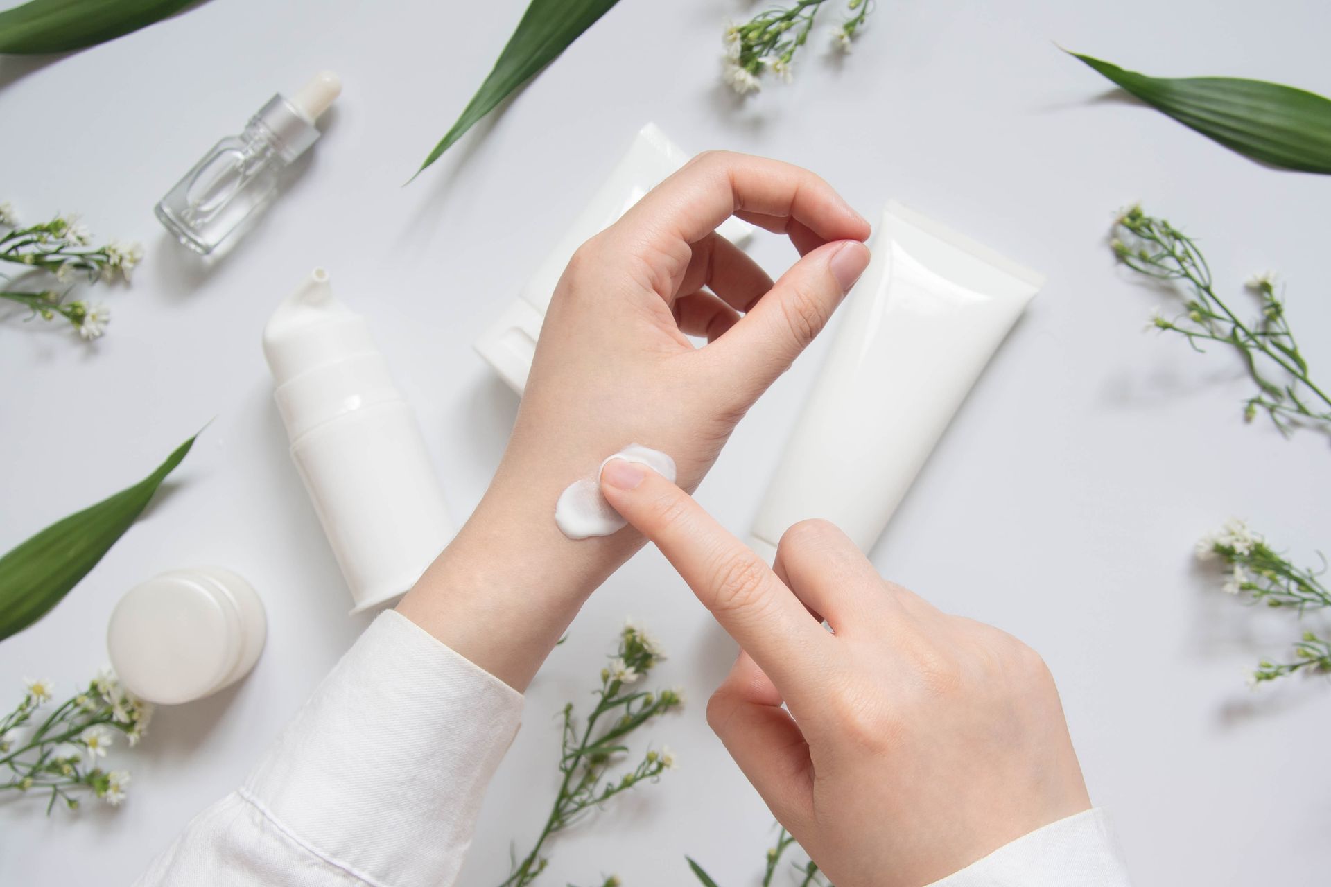 Des mains appliquant une lotion blanche sur un poignet, à côté de flacons de soins pour la peau et de feuilles vertes sur un fond clair.