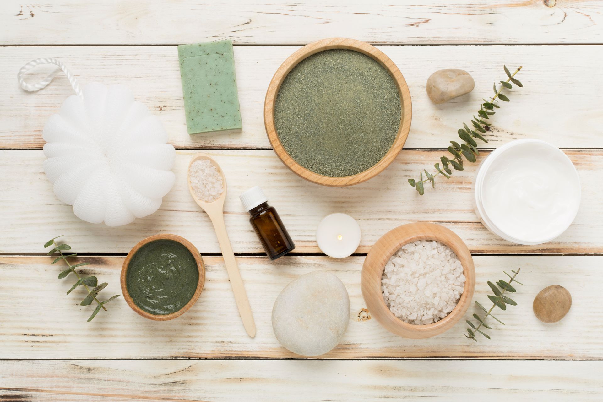 Assortiment d'ingrédients et d'outils naturels pour les soins de la peau disposés sur une surface en bois blanc, comprenant des bols, des pinceaux, du savon et des herbes.