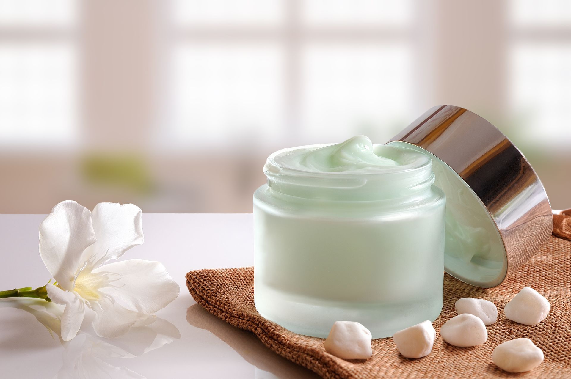 Pot de crème pour le visage vert menthe avec couvercle ouvert, fleur blanche et galets lisses sur un plateau de spa