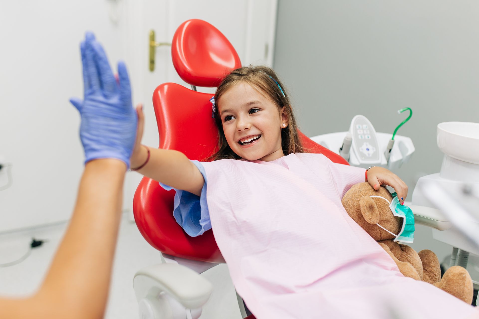 Un enfant souriant, assis sur un fauteuil de dentiste, fait un clap de la main avec une autre gantée, avec un ours en peluche et du matériel dentaire à proximité.