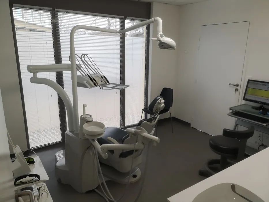 Salle d'examen dentaire avec fauteuil, éclairage au plafond, écran et stores.